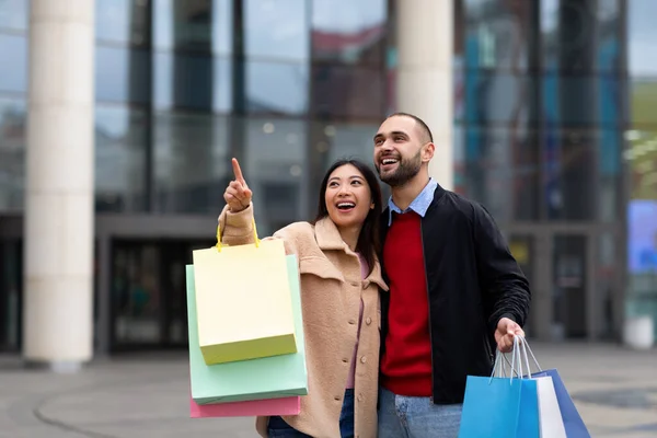 Multiraciaal stel loopt door straat met boodschappentassen, wijst naar nieuwe winkel of advertentie, gaat samen winkelen — Stockfoto