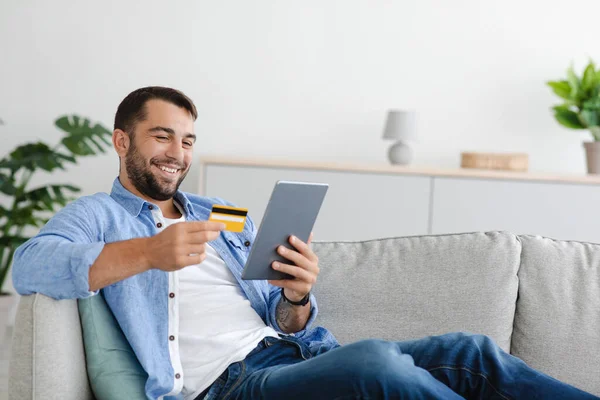 Alegre alegre guapo maduro caucásico chico con barba sentado en sofá celebrar tarjeta de crédito y tableta, compras en línea — Foto de Stock