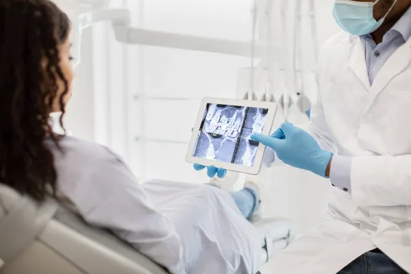 Médico dentista preto mostrando raios-X dentes em comprimido digital para paciente feminino — Fotografia de Stock