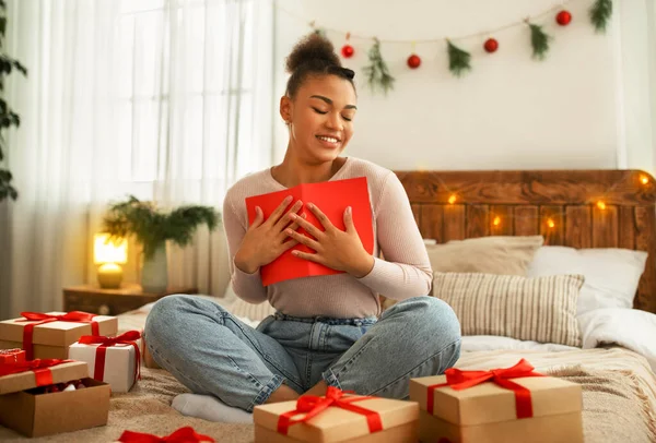 Felice signora afro-americana che celebra il nuovo anno seduto sul letto e abbraccia il biglietto di Natale ad occhi chiusi — Foto Stock