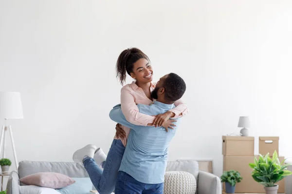 Leende ung afrikansk amerikansk make kramar och lyfter sin fru i ny lägenhet med pappkartonger — Stockfoto