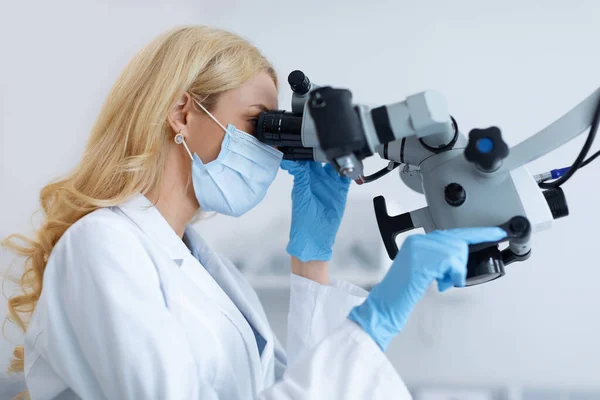 Blond kvinna i ansiktet mask stomatolog tittar på dentalt mikroskop — Stockfoto