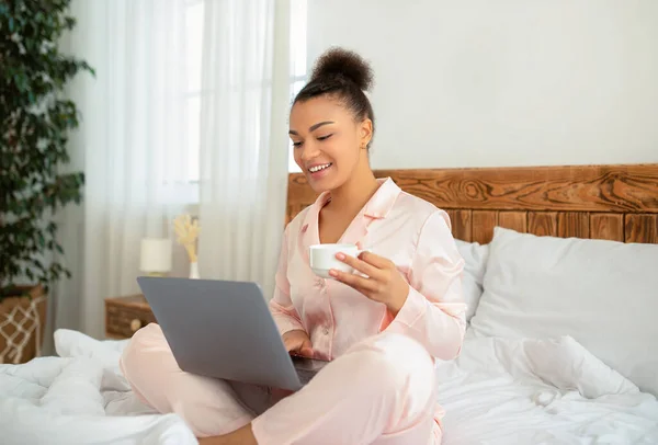 Trabajo independiente. Feliz dama afroamericana sentada en la cama con portátil y taza de café, quedarse en casa, copiar espacio — Foto de Stock