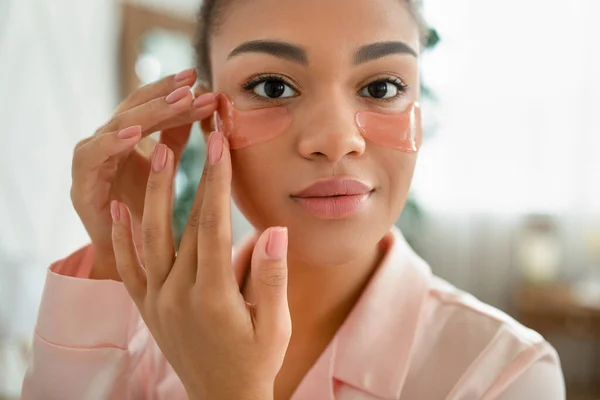 Pielęgnacja twarzy. Afroamerykanka stosująca plastry kosmetyczne pod oczami nawilżające skórę i uśmiechnięta do aparatu — Zdjęcie stockowe