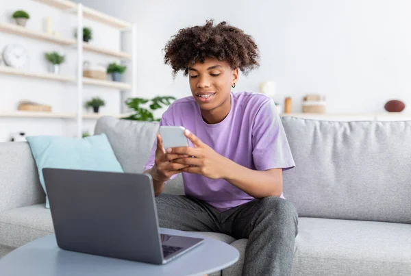 Jovem feliz adolescente preto em desgaste casual usando smartphone e laptop, estudando on-line de casa — Fotografia de Stock