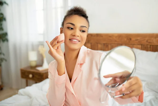 Senhora americana muito africana aplicando fundação cosmética com aplicador, segurando espelho, sentado na cama no quarto — Fotografia de Stock