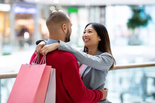 Preciosa pareja joven y diversa con bolsas de compras abrazándose y mirándose dentro de un gran supermercado —  Fotos de Stock