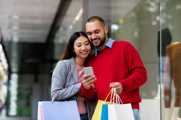 Веселая молодая межрасовая пара с подарочными пакетами и мобильным телефоном, заказывающая товары онлайн возле торгового центра — стоковое фото