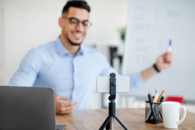 Neşeli Arap erkek öğretmen çevrimiçi İngilizce dersi veriyor, akıllı telefonu tripod ve dizüstü bilgisayarda kullanıyor, seçici odak noktası