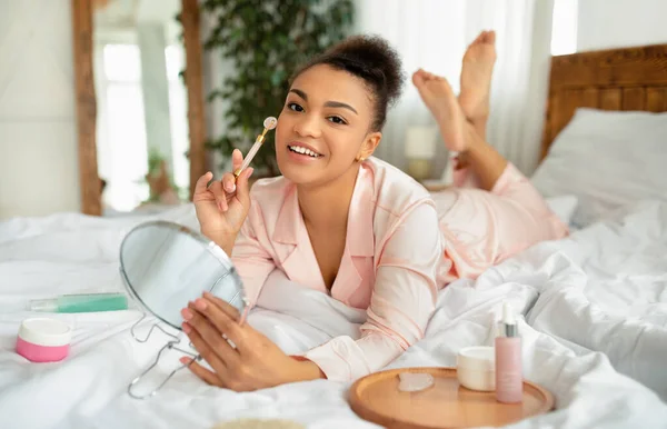 Afrika Amerika wanita dalam setelan malam melakukan pijat wajah menggunakan rol, berbaring di tempat tidur dengan produk kecantikan dan cermin — Stok Foto