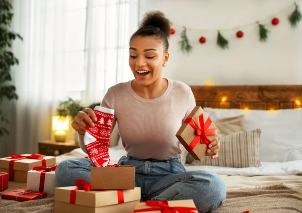 Festeggiamenti di Capodanno. Eccitato signora afroamericana che riceve regalo di Natale e tenendo calzini di Natale, seduto sul letto — Foto Stock