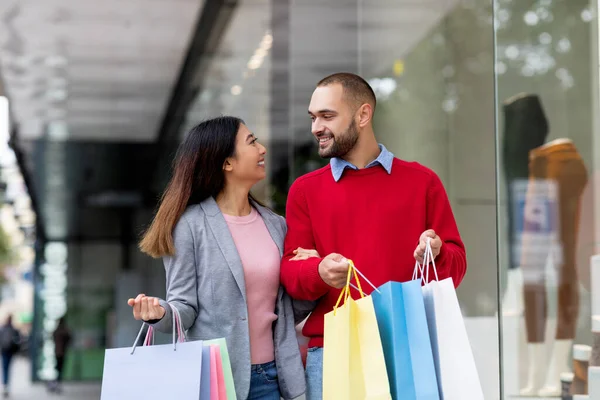 Portret van lachende jonge interraciale paar met boodschappentassen kijken naar elkaar in winkelcentrum — Stockfoto