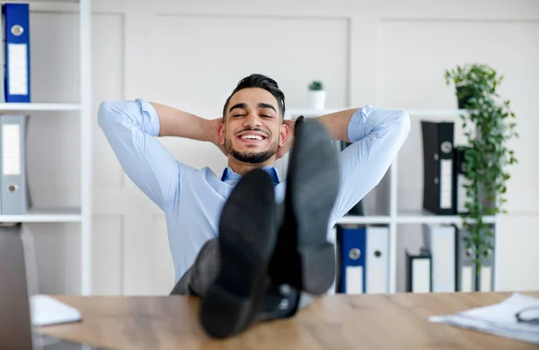Счастливый араб тысячелетия расслабляется после успешной работы, ставя ноги на стол в современном офисе — стоковое фото