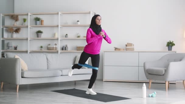 Молода позитивна мусульманка займається еластичною гумкою, роблячи спортивну практику для ніг з додатковим навантаженням вдома — стокове відео