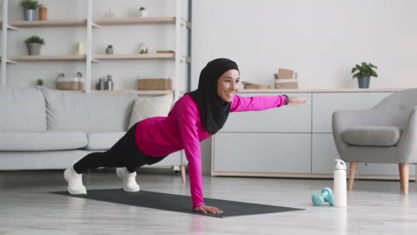 Entrenamiento de fuerza en casa. Joven mujer musulmana activa en hijab practicando ejercicio dinámico de tablón, levantando la mano en el suelo — Vídeos de Stock