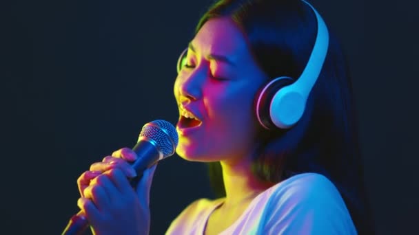 Penyanyi yang riang. Wanita asia muda di headphone nirkabel menyanyikan lagu favorit di mikrofon di lampu neon terang — Stok Video