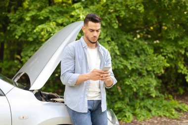 Kafası karışmış orta doğulu sürücü bozuk arabanın yanında duruyor, cep telefonu kullanıyor.