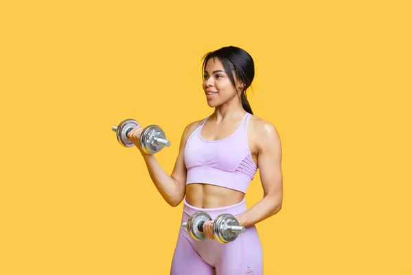 Concepto de entrenamiento de fuerza. Ajuste mujer afroamericana haciendo ejercicio con dos pesas, fondo de estudio amarillo —  Fotos de Stock