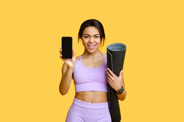 Conceito de aplicativo de esportes. Happy fit preto senhora segurando smartphone com tela vazia e tapete de ioga, fundo amarelo — Fotografia de Stock