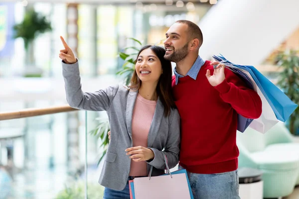 Faire du shopping ensemble. Millennial interracial couple achetant des marchandises au supermarché, tenant des sacs à provisions, pointant de côté — Photo