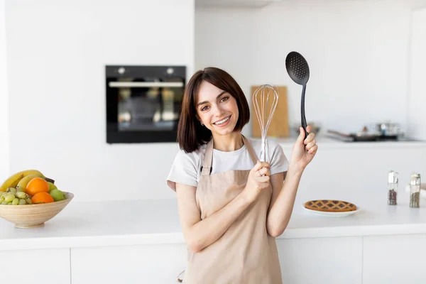Porträtt av glad dam som bär förkläde och poserar i köket håller köksredskap i händerna och ler mot kameran — Stockfoto