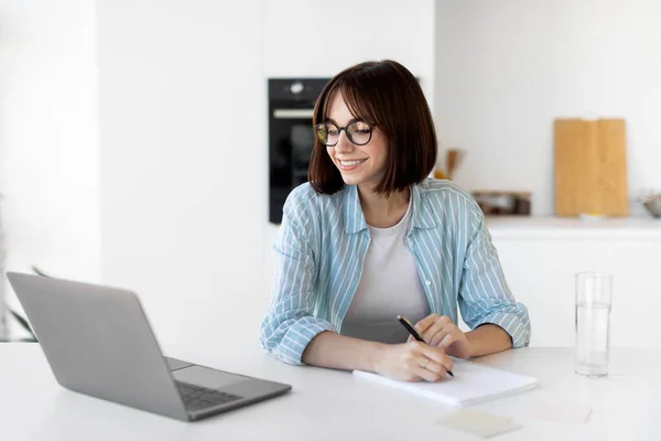 Jovem freelancer mulher trabalhando com laptop na cozinha e escrever informações enquanto está sentado na cozinha — Fotografia de Stock