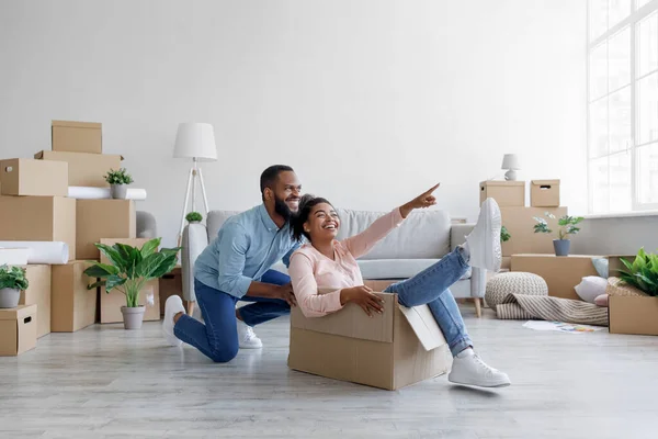 Szczęśliwy czarny mężczyzna pchający śmiejącą się żonę w pudełku kartonowym. Energiczna kobieta siedzi w kartonowym pojemniku, baw się dobrze — Zdjęcie stockowe