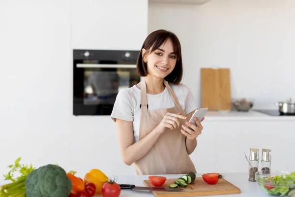 Glad tusenårig kvinna med smartphone och söker nytt recept, står i köket och ler mot kameran — Stockfoto