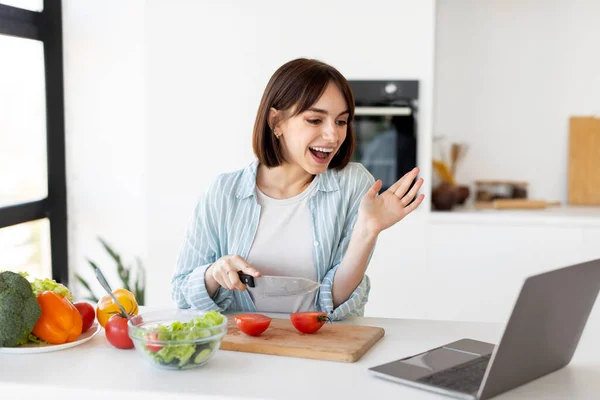 집에서 음식을 준비 한다. 야채를 자르고 샐러드를 준비하는 긍정적 인 아가씨, 노트북을 통해 비디오 채팅을 하는 모습 — 스톡 사진