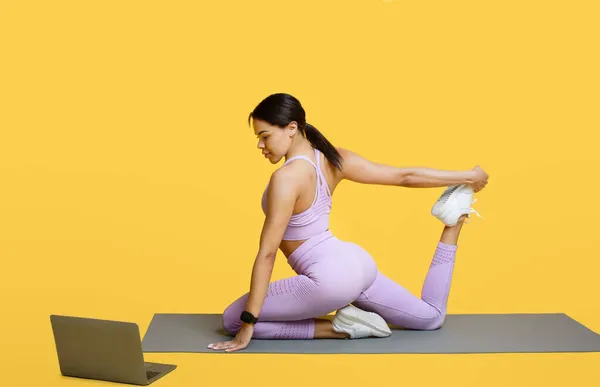 健康在线。年轻的黑人妇女伸展身体，坐在笔记本电脑前的瑜伽垫上，在黄色的背景上锻炼 — 图库照片