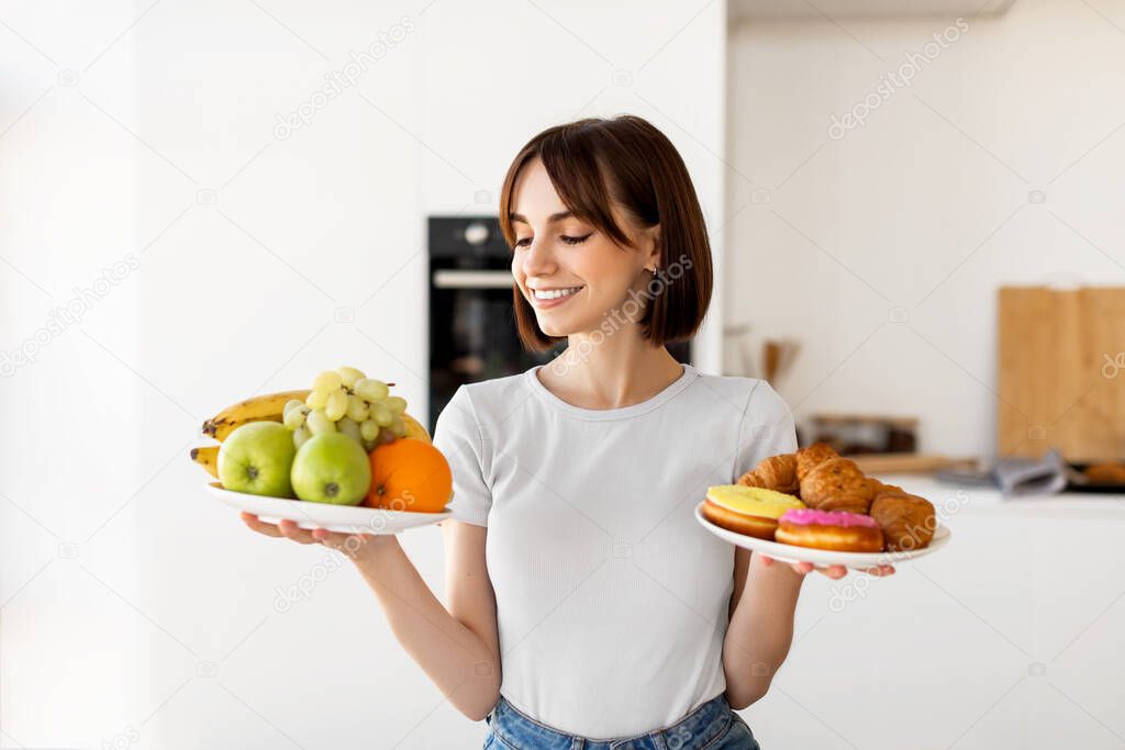 Choosing between healthy or unhealthy food. Young woman holding plates with fruits and sweets, standing in kitchen
