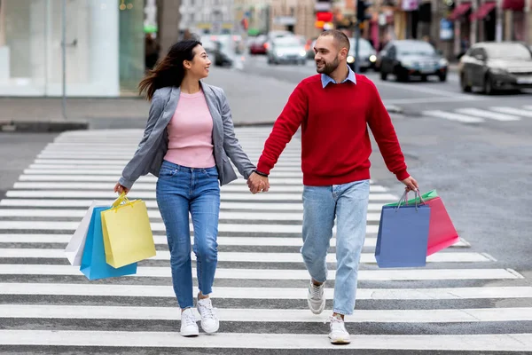 Full length portret van jong multiraciaal paar met shopper tassen oversteken straat, hand in hand buiten — Stockfoto