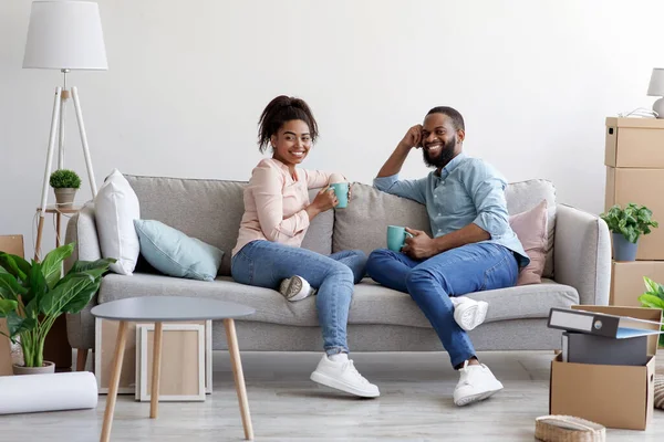 Sorrindo jovem afro-americano homem e mulher relaxar no sofá, beber café no quarto com caixas de papelão — Fotografia de Stock