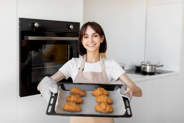 Glad ung hemmafru som håller bricka med färska bakade croissanter, står i köket och ler mot kameran — Stockfoto