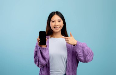 Akıllı telefonu boş ekranla gösteren mutlu Asyalı kadın, yeni mobil uygulama veya web sitesi öneriyor, mavi arkaplan