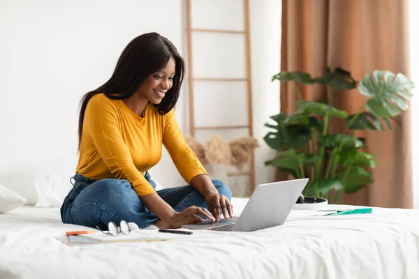 Svart ung kvinna med hjälp av laptop skriva sitta i sovrummet inomhus — Stockfoto