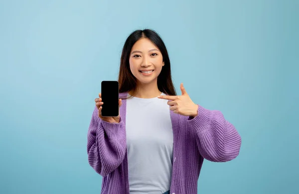 Felice signora asiatica che punta a smartphone con schermo vuoto, raccomandando nuova applicazione mobile o sito web, sfondo blu — Foto Stock