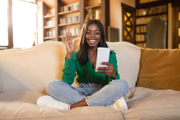Felice donna afroamericana con videochiamata su smartphone, agitando la fotocamera mentre si rilassa sul divano di casa — Foto Stock