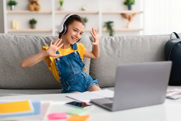 Chica sentada en el sofá, teniendo videollamada con auriculares saludando — Foto de Stock