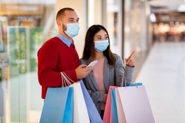 Yüz maskesi takan, alışveriş merkezinde alışveriş torbalarıyla yürüyen akıllı telefondan alışveriş listesini kontrol eden çok ırklı genç bir çift.