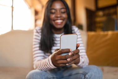 Akıllı telefon ağı olan mutlu siyah kadın, e-postaları kontrol ediyor, sosyal ağda paylaşıyor, evdeki kanepeye mesaj yolluyor.