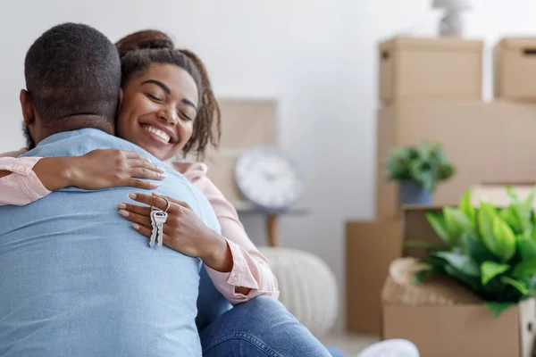 Satisfied young black family hugging, hold key and enjoy buying real estate in room with cardboard boxes and plants — Stock Photo, Image