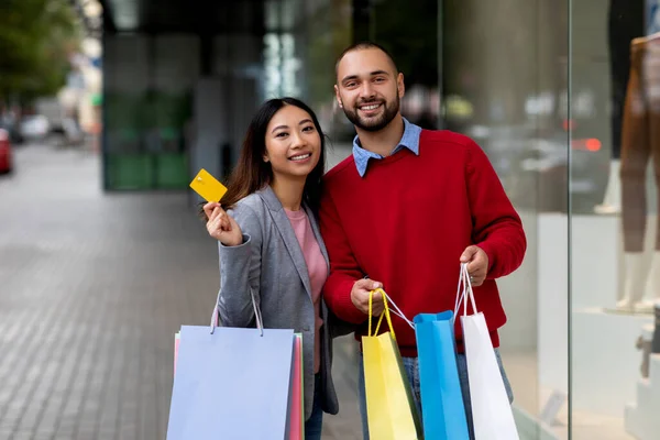 Para wielonarodowa pozująca w pobliżu centrum handlowego, trzymająca kartę kredytową i torby na zakupy, promująca płatności zbliżeniowe — Zdjęcie stockowe