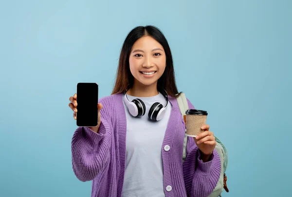 Возбужденная азиатская студентка показывает смартфон с чистым экраном, позирует с рюкзаком и кофе, макет — стоковое фото