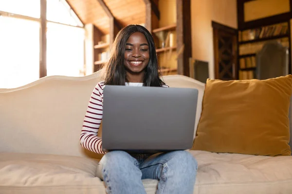 Online jobb och utbildning. Glädjande ung svart kvinna sitter på soffan med bärbar dator, arbetar eller lär hemifrån — Stockfoto