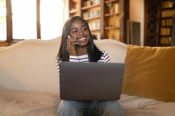 Online business meeting. Happy young black woman using laptop for video conference, calling on moile phone at home