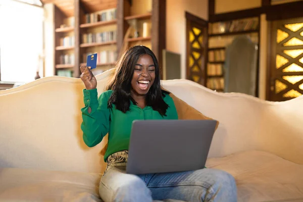 Excited young black lady with credit card and laptop shouting OMG, winning in online lottery, shopping on sale from home