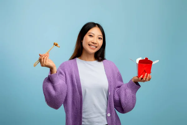 Entrega de comida, concepto de comida rápida. Asiática señora comiendo fideos con palillos, sosteniendo caja de papel sobre fondo azul —  Fotos de Stock