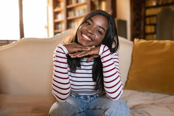 Retrato de una joven afroamericana apoyada en sus manos y sonriendo, mirando a la cámara, sentada en el sofá en casa — Foto de Stock