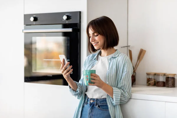 Redes sociales y chat por la mañana. Feliz joven bebiendo café y usando smartphone, de pie en la cocina — Foto de Stock